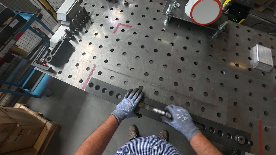 Zicht door de ogen van een operator: twee handen in werkhandschoenen leggen een staaf op een lastafel naast een cobot.